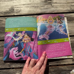 Pode incluir: Livro aberto com ilustra&ccedil;&otilde;es coloridas de sereias e um golfinho rosa. As ilustra&ccedil;&otilde;es apresentam sereias com cabelos longos e caudas de sereia em um cen&aacute;rio de fantasia. O livro est&aacute; aberto em uma superf&iacute;cie de madeira.
