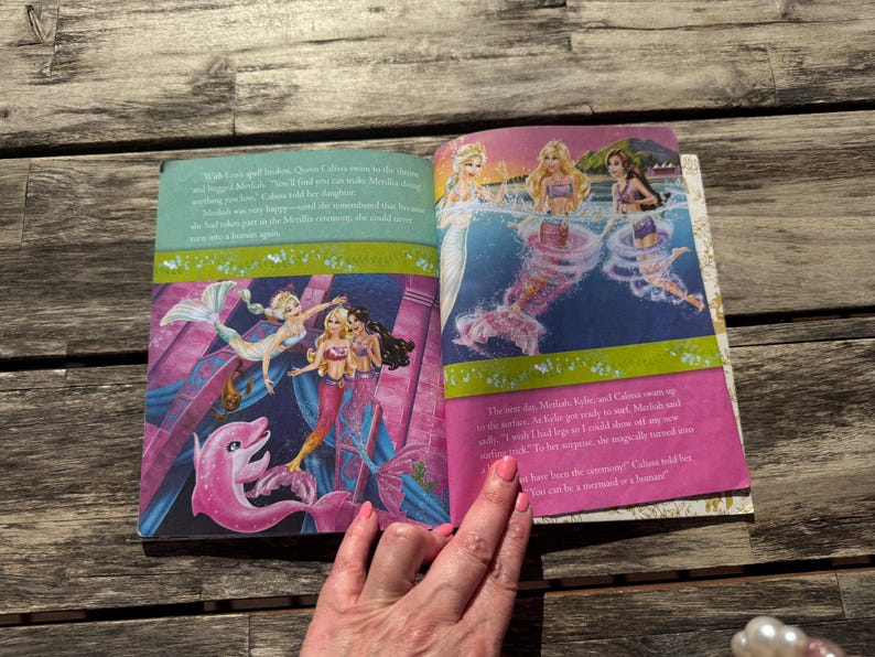 Pode incluir: Livro aberto com ilustra&ccedil;&otilde;es coloridas de sereias e um golfinho rosa. As ilustra&ccedil;&otilde;es apresentam sereias com cabelos longos e caudas de sereia em um cen&aacute;rio de fantasia. O livro est&aacute; aberto em uma superf&iacute;cie de madeira.