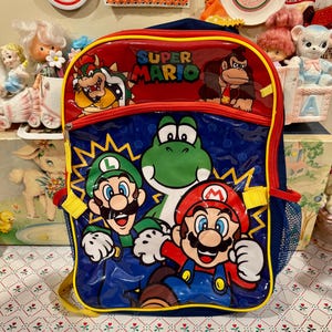 May include: A blue and red Super Mario backpack featuring a glossy front panel with graphics of Mario, Luigi, Yoshi, Bowser, and Donkey Kong. The bag includes a zippered main compartment, a smaller front pocket, and blue mesh side pockets.