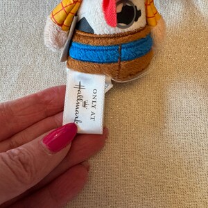 May include: A small plush toy depicting a cartoon cowboy, with a brown hat, yellow shirt, and blue jeans. The toy has a red neckerchief and a tag that reads "ONLY AT Hallmark".