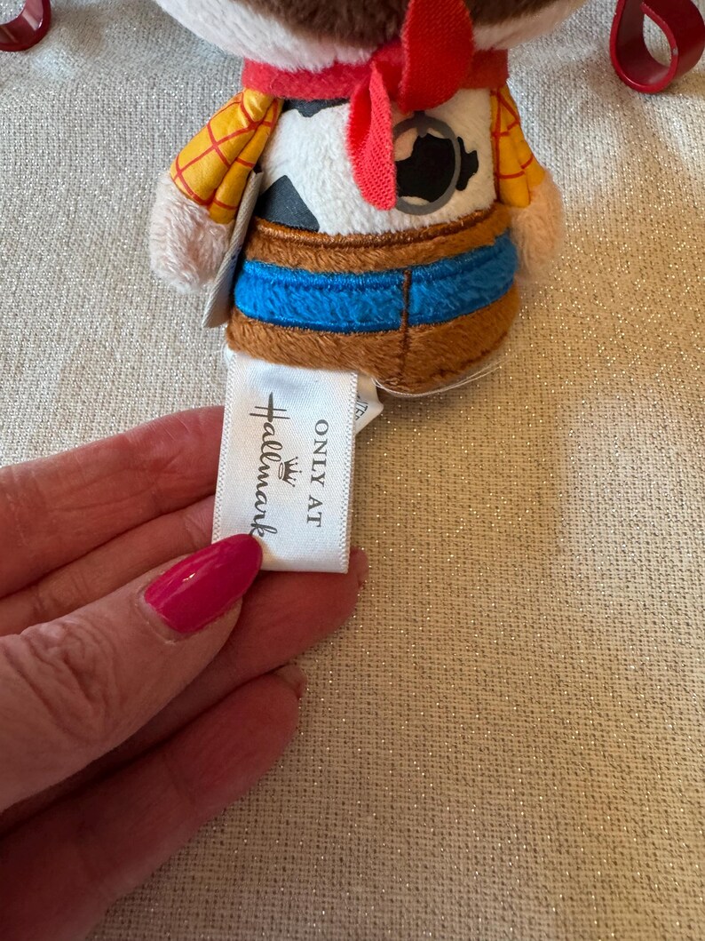 May include: A small plush toy depicting a cartoon cowboy, with a brown hat, yellow shirt, and blue jeans. The toy has a red neckerchief and a tag that reads "ONLY AT Hallmark".