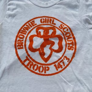 May include: A white short-sleeved t-shirt featuring a circular orange graphic on the chest. The graphic displays the text "BROWNIE GIRL SCOUTS" and "TROOP 1473" surrounding a central clover-shaped emblem with a stylized figure in the middle.
