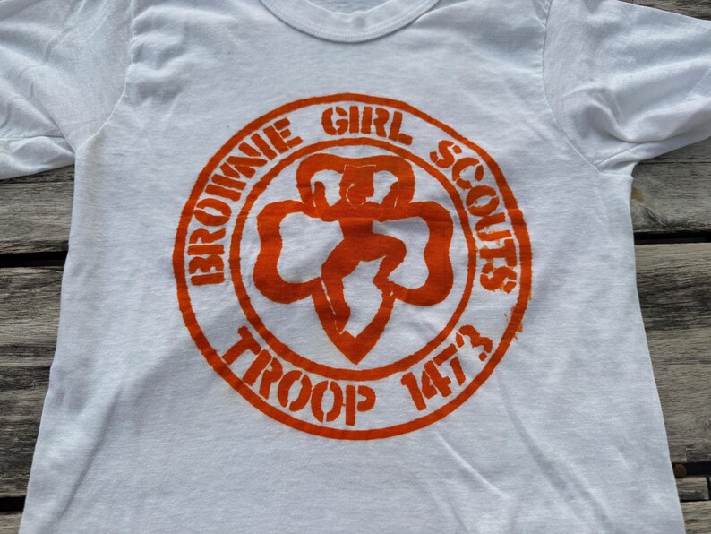 May include: A white short-sleeved t-shirt featuring a circular orange graphic on the chest. The graphic displays the text "BROWNIE GIRL SCOUTS" and "TROOP 1473" surrounding a central clover-shaped emblem with a stylized figure in the middle.