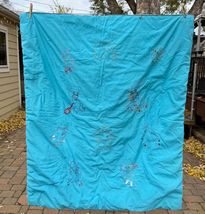 May include: A turquoise quilt with embroidered teddy bears and other whimsical figures. The quilt is hanging outside on a clothesline. The embroidery is in various colors, including red, yellow, and blue.