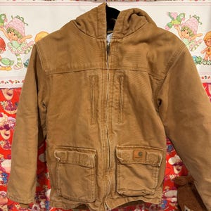 Puede incluir: Chaqueta con capucha marr&oacute;n con cierre de cremallera, dos bolsillos en el pecho y dos bolsillos inferiores. La chaqueta tiene el logotipo de Carhartt en el bolsillo inferior derecho. La chaqueta est&aacute; colgada en una percha negra.