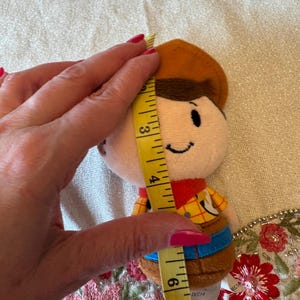 May include: A plush toy of a cartoon cowboy character, approximately 15 cm tall. The toy has a brown hat, a yellow shirt with a plaid pattern, and blue pants. The toy is being measured with a yellow measuring tape.