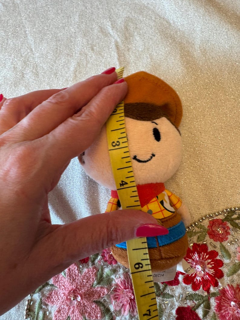 May include: A plush toy of a cartoon cowboy character, approximately 15 cm tall. The toy has a brown hat, a yellow shirt with a plaid pattern, and blue pants. The toy is being measured with a yellow measuring tape.