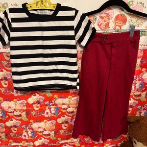 May include: A black and white striped t-shirt and a pair of red pants. The t-shirt has short sleeves and a round neckline. The pants are high-waisted with a button closure. The clothing is displayed on a colorful background.