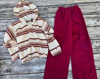 Puede incluir: Una sudadera con capucha y manga larga con rayas horizontales en crema, negro y rojo óxido, combinada con pantalones anchos de color magenta intenso con cintura elástica. La sudadera tiene un cuello en V y una capucha con cordón.