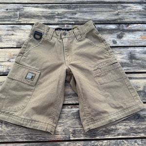 May include: Khaki-colored cargo shorts with multiple pockets and a metal D-ring. The shorts have a rectangular patch on the front pocket. The shorts are made of a durable fabric and are suitable for outdoor activities.