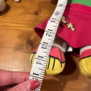 May include: A plush toy dog wearing a red, green, and yellow outfit. The dog is holding a measuring tape that reads 14 inches.