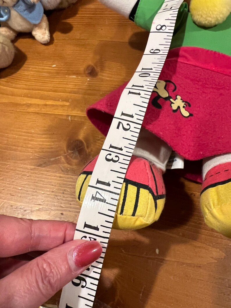 May include: A plush toy dog wearing a red, green, and yellow outfit. The dog is holding a measuring tape that reads 14 inches.