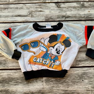 May include: A white long-sleeve sweatshirt featuring a graphic of Mickey Mouse wearing sunglasses with the text CATCH IT. The garment has light blue sleeves with black and red stripes, a black crew neckline, black cuffs, and a black waistband.