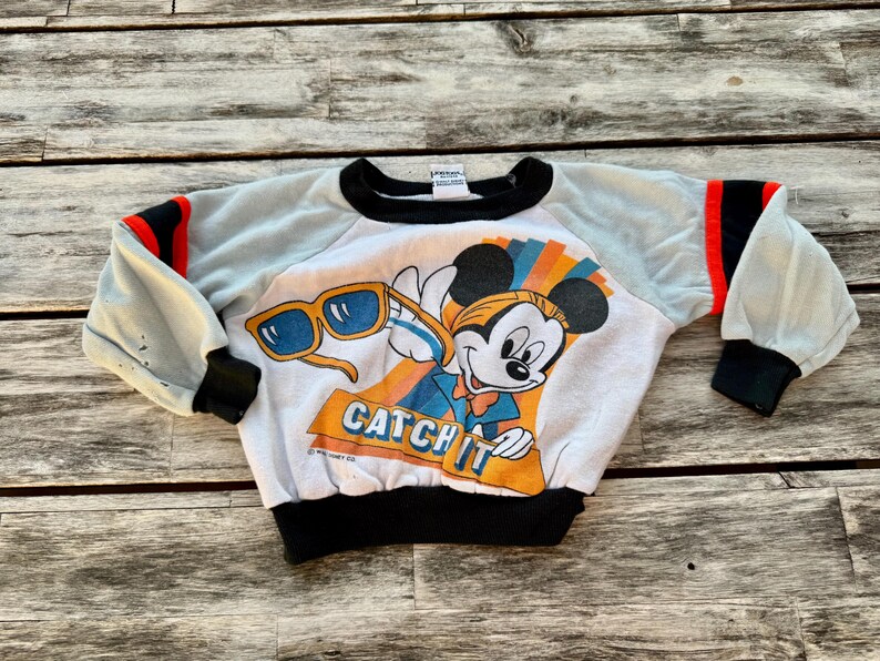 May include: A white long-sleeve sweatshirt featuring a graphic of Mickey Mouse wearing sunglasses with the text CATCH IT. The garment has light blue sleeves with black and red stripes, a black crew neckline, black cuffs, and a black waistband.
