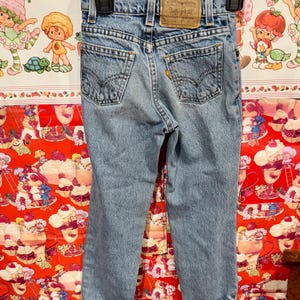 May include: Light blue denim jeans with classic five-pocket styling, featuring a brown leather Levi Strauss & Co. patch on the back. The jeans are hanging on a black hanger, showcasing the back pockets and stitching details.