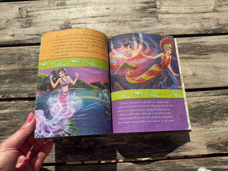 Pode incluir: Livro aberto com ilustra&ccedil;&otilde;es coloridas de sereias. Uma sereia est&aacute; na &aacute;gua, a outra numa rocha. O texto descreve uma aventura de sereia. O livro est&aacute; sobre uma superf&iacute;cie de madeira.