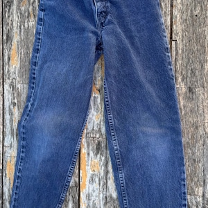 May include: A pair of blue denim jeans with a classic five-pocket design. The jeans have a button and zipper closure, belt loops, and are made of a sturdy denim fabric. The jeans are displayed on a weathered wooden surface.