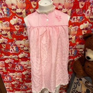 May include: A vintage pink nightgown with a white lace trim and a small butterfly embroidery. The sleeveless gown features a ruffled collar and a gingham yoke. The nightgown is displayed on a mannequin.
