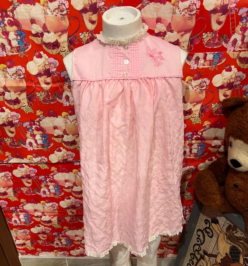 May include: A vintage pink nightgown with a white lace trim and a small butterfly embroidery. The sleeveless gown features a ruffled collar and a gingham yoke. The nightgown is displayed on a mannequin.