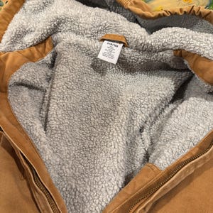 Puede incluir: Primer plano de una chaqueta con capucha marr&oacute;n con un forro interior gris y difuso. La chaqueta tiene cierre de cremallera y una peque&ntilde;a etiqueta con texto. La chaqueta parece estar hecha de un material duradero.