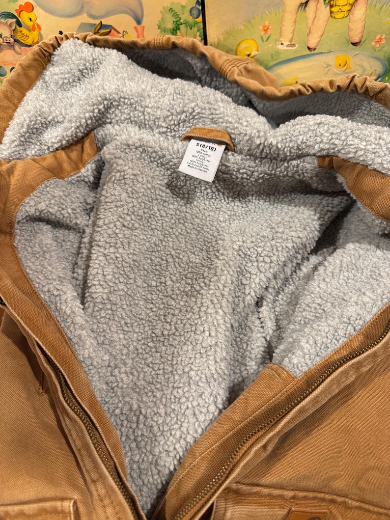 Puede incluir: Primer plano de una chaqueta con capucha marr&oacute;n con un forro interior gris y difuso. La chaqueta tiene cierre de cremallera y una peque&ntilde;a etiqueta con texto. La chaqueta parece estar hecha de un material duradero.
