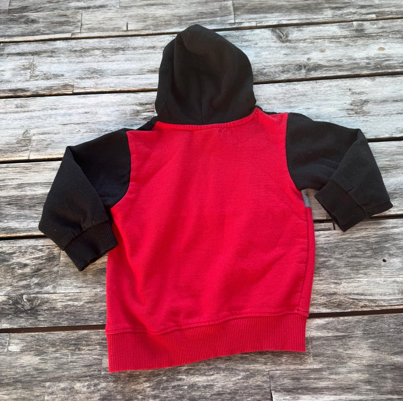 May include: A long-sleeve hooded sweatshirt featuring a vibrant red main body contrasted by solid black sleeves and a black hood. The garment includes ribbed cuffs at the wrists and a ribbed hem at the bottom, showcasing a two-tone color block design.