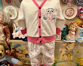 Puede incluir: Un conjunto de ropa de bebé vintage con una rebeca blanca con ribetes rosas y botones negros, combinada con pantalones a juego. El conjunto tiene un diseño de oso bordado en el pecho y un patrón de oso repetido en los pantalones. El conjunto es de un material de rizo suave.