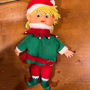 May include: A vintage elf doll with a red hat, green shirt, and red pants. The doll has yellow hair, a red collar, and green shoes. The belt is red with a gold buckle. The doll is a festive holiday decoration.