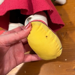 May include: Close-up of a yellow fabric shoe with a black outline. The shoe is part of a plush toy doll wearing a red skirt and green top.