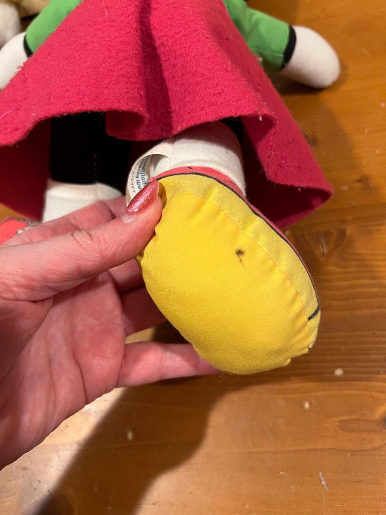 May include: Close-up of a yellow fabric shoe with a black outline. The shoe is part of a plush toy doll wearing a red skirt and green top.