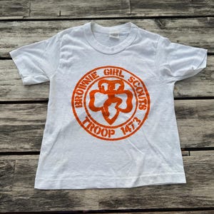 May include: A white short-sleeve t-shirt featuring an orange circular graphic on the chest. The graphic displays the text "BROWNIE GIRL SCOUTS TROOP 1473" surrounding a central clover-shaped emblem with a stylized figure in the middle.