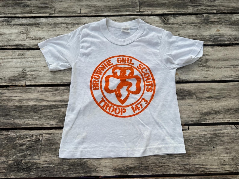 May include: A white short-sleeve t-shirt featuring an orange circular graphic on the chest. The graphic displays the text "BROWNIE GIRL SCOUTS TROOP 1473" surrounding a central clover-shaped emblem with a stylized figure in the middle.