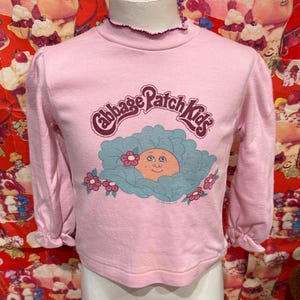 May include: A pink long-sleeved shirt featuring the text "Cabbage Patch Kids" in a purple and white design. The shirt has a ruffled collar and cuffs, and a graphic of a baby's face in a cabbage.