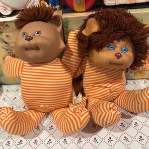 May include: Two plush dolls with tan skin and round faces wearing orange and white striped long-sleeved outfits. The doll on the left has short brown hair and gray eyes, while the doll on the right has shaggy brown hair and bright blue eyes.