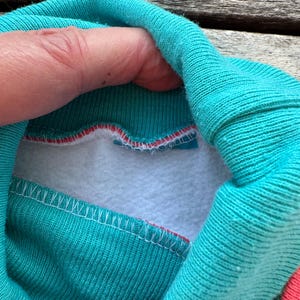 Op de afbeelding: Een groenblauwe opstaande kraag met een witte fleece voering aan de binnenzijde. De kraag is aan de buitenkant gemaakt van geribbelde gebreide stof en bevat een dunne rode accentlijn langs de binnennaad waar de kraag de fleece voering raakt.