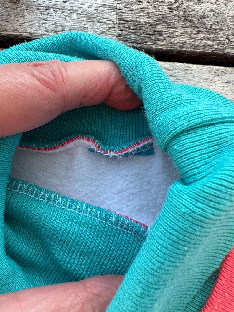 Op de afbeelding: Een groenblauwe opstaande kraag met een witte fleece voering aan de binnenzijde. De kraag is aan de buitenkant gemaakt van geribbelde gebreide stof en bevat een dunne rode accentlijn langs de binnennaad waar de kraag de fleece voering raakt.