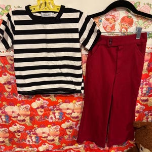 May include: A black and white striped t-shirt and a pair of red pants hanging on a hanger. The t-shirt has short sleeves and a round neckline. The pants are high-waisted with a button closure.