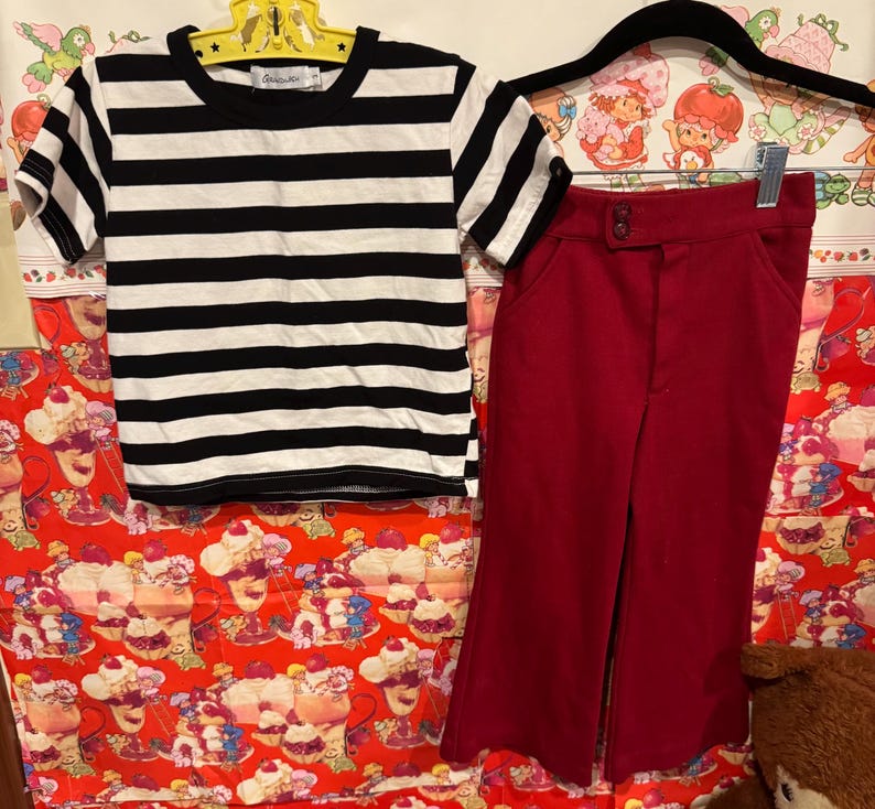 May include: A black and white striped t-shirt and a pair of red pants hanging on a hanger. The t-shirt has short sleeves and a round neckline. The pants are high-waisted with a button closure.