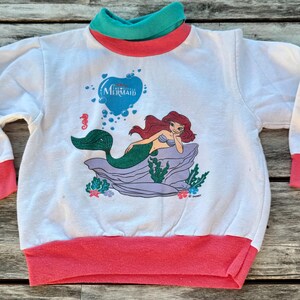 Op de afbeelding: Een wit sweatshirt met lange mouwen en een afbeelding van de kleine zeemeermin Ariel op een rots. Het kledingstuk heeft een blauwgroene opstaande kraag, koraalroze mouwboorden en een koraalroze zoom. De tekst The Little Mermaid staat erboven.