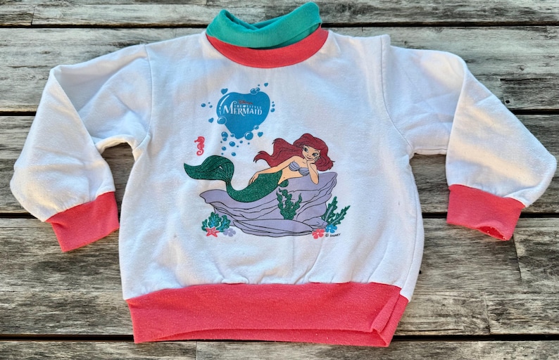 Op de afbeelding: Een wit sweatshirt met lange mouwen en een afbeelding van de kleine zeemeermin Ariel op een rots. Het kledingstuk heeft een blauwgroene opstaande kraag, koraalroze mouwboorden en een koraalroze zoom. De tekst The Little Mermaid staat erboven.
