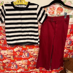 May include: A black and white striped t-shirt and a pair of red pants. The t-shirt has short sleeves and a round neckline. The pants are high-waisted with a button closure. The clothing is displayed on a colorful background.