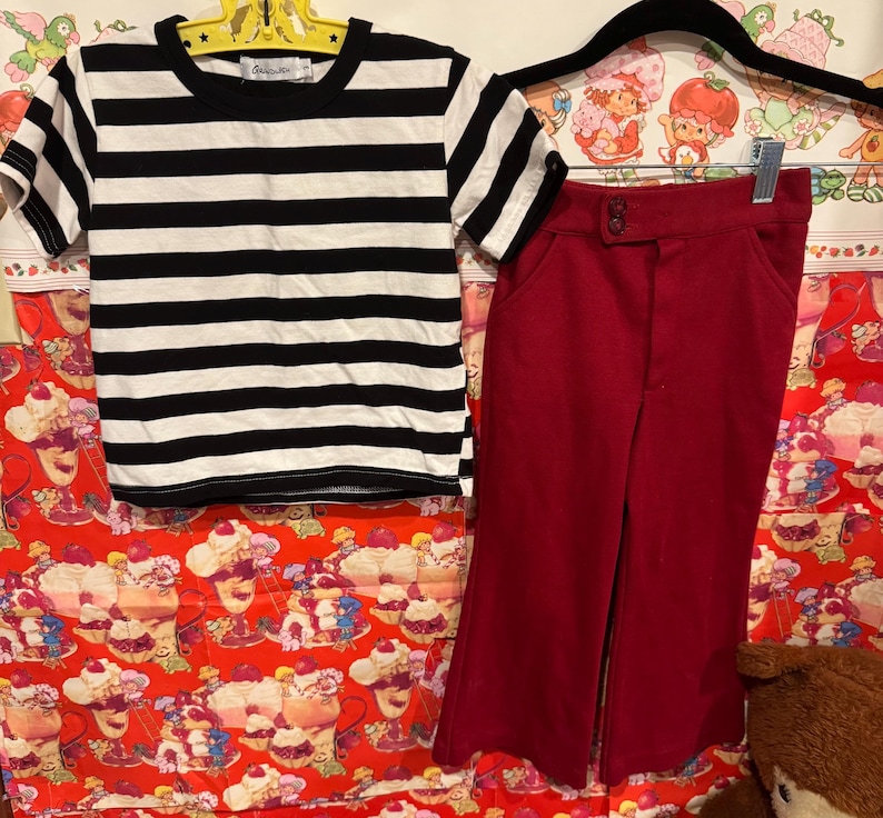 May include: A black and white striped t-shirt and a pair of red pants. The t-shirt has short sleeves and a round neckline. The pants are high-waisted with a button closure. The clothing is displayed on a colorful background.