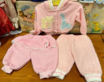 Puede incluir: Un conjunto de ropa de bebé en rosa suave, con una chaqueta con capucha con aplicaciones de elefante y jirafa, y dos pantalones. Uno tiene una cintura con volantes con ribete de encaje, y el otro tiene un patrón acolchado. El conjunto está diseñado para bebés.
