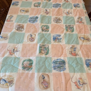 May include: A quilted blanket with a patchwork design featuring alternating squares of pink and light blue. Each square contains a different illustration of animals, including mice and ducks. The blanket is placed on a wooden table.