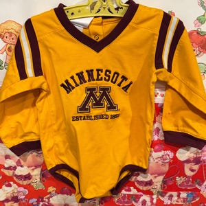 May include: A yellow baby bodysuit with long sleeves. The bodysuit has a maroon V-neck collar and cuffs, with white stripes on the sleeves. The word "MINNESOTA" is arched above a maroon "M" logo, with "ESTABLISHED 1851" below.