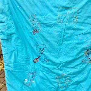 May include: A turquoise baby quilt with embroidered teddy bear designs. The bears are in various poses, some holding toys. The embroidery is in shades of pink, red, and yellow. The quilt has a quilted texture.