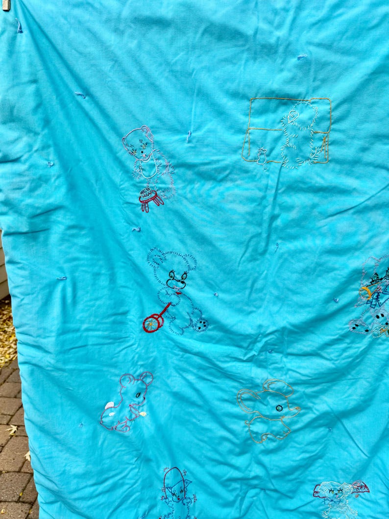 May include: A turquoise baby quilt with embroidered teddy bear designs. The bears are in various poses, some holding toys. The embroidery is in shades of pink, red, and yellow. The quilt has a quilted texture.