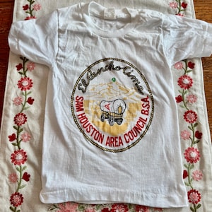 May include: White t-shirt featuring a circular emblem with the text "El Rancho Cima" and "Sam Houston Area Council B.S.A." The emblem includes a covered wagon illustration and mountain scene. The shirt is short-sleeved.