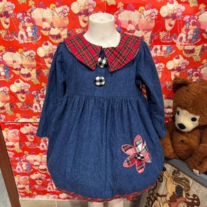 May include: A blue denim dress with long sleeves, a red tartan collar, and a flower appliqué. The dress has a red tartan trim at the hem and is fastened with black and white buttons. A teddy bear is in the background.