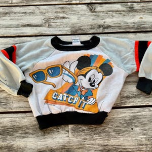 May include: A white long-sleeve sweatshirt featuring a graphic of Mickey Mouse holding yellow sunglasses with the text CATCH IT. The garment has light gray sleeves with black and red stripes at the shoulders, black cuffs, and a black crew neckline.